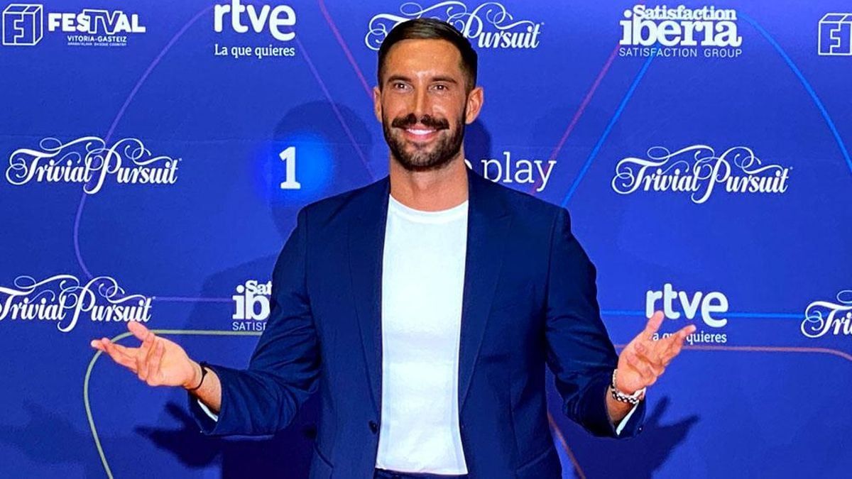 Hombre sonriendo en evento de RTVE con fondo de logos y promoción de 'Trivial Pursuit'.