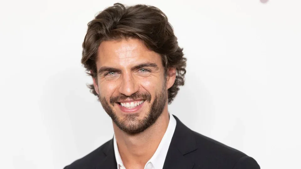 Maxi Iglesias sonríe en primer plano, con traje oscuro y camisa blanca, ante el photocall de un evento